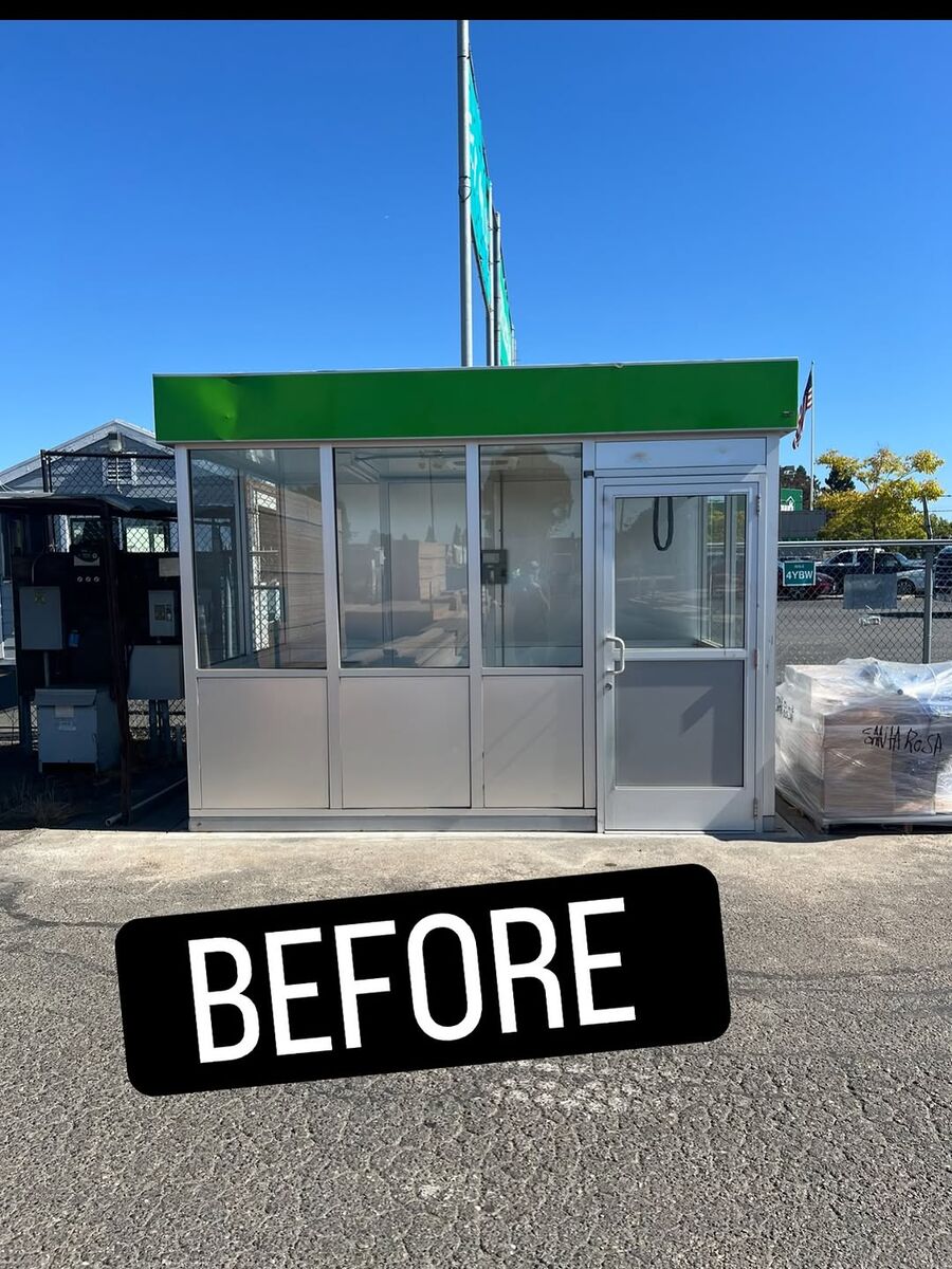 Small glass-walled attendant booth with a green fascia photographed before mirror window film was applied, a BEFORE placard in front of it