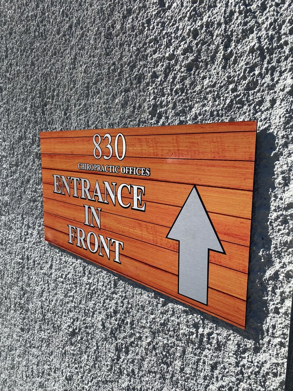 Wood-grain directional sign reading 830 Chiropractic Offices, Entrance in Front, with a raised silver arrow pointing up, mounted on a heavily textured grey stucco wall