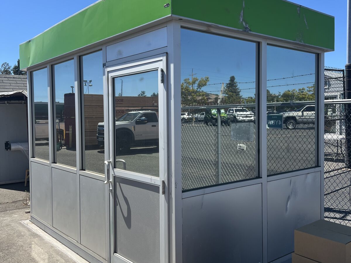 Small glass-walled attendant booth with a green fascia, its windows finished in mirror film that reflects the parking lot