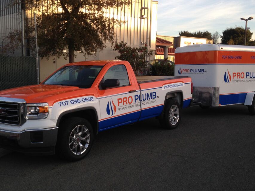 White GMC Sierra pickup with orange and blue Pro Plumb graphics, towing an enclosed cargo trailer carrying the same graphics