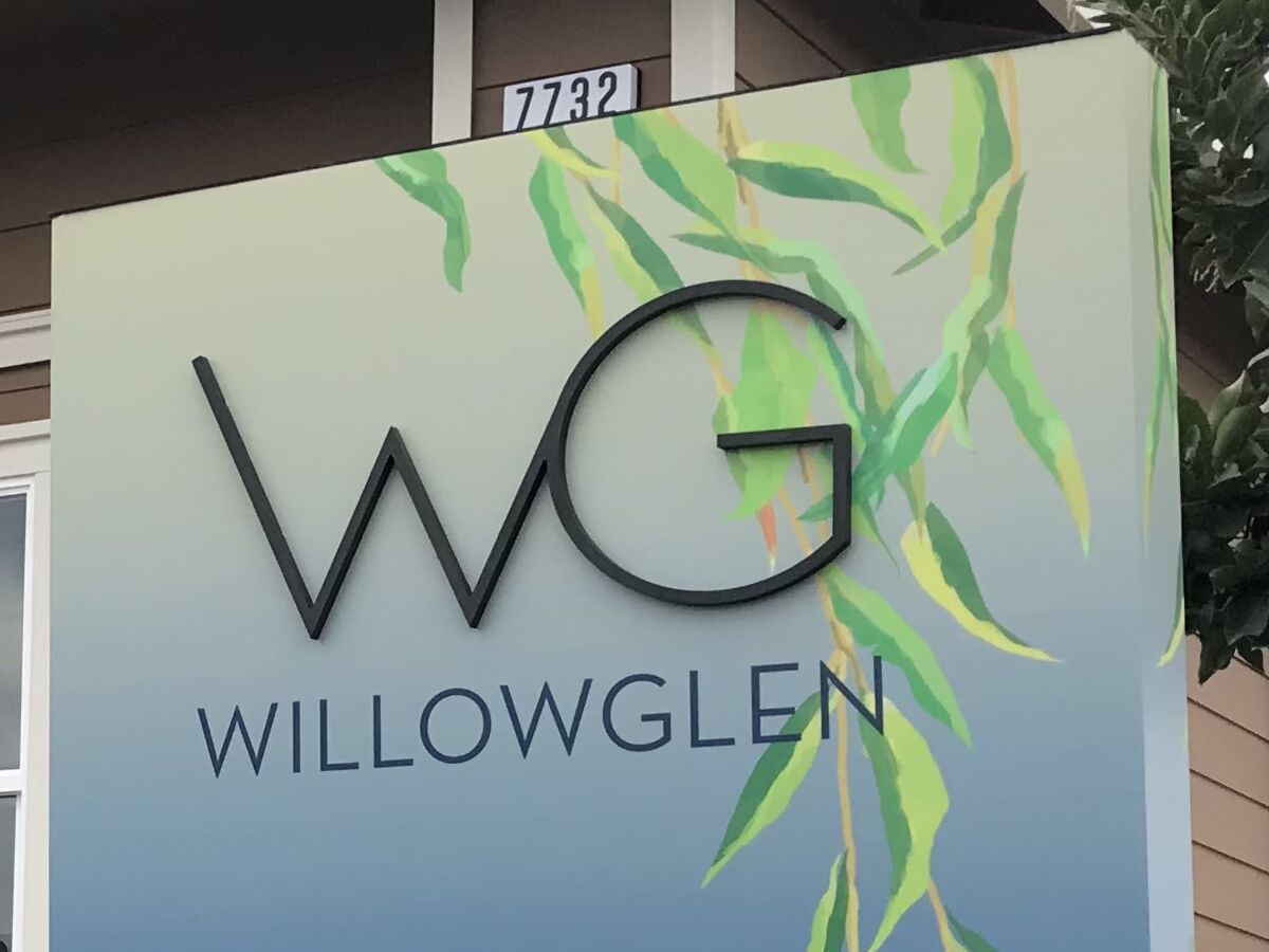 Close view of the WillowGlen sales-office sign: dimensional WG letters standing off a cream-to-blue gradient face, the word WillowGlen beneath them, and a painted willow branch trailing down the right-hand side