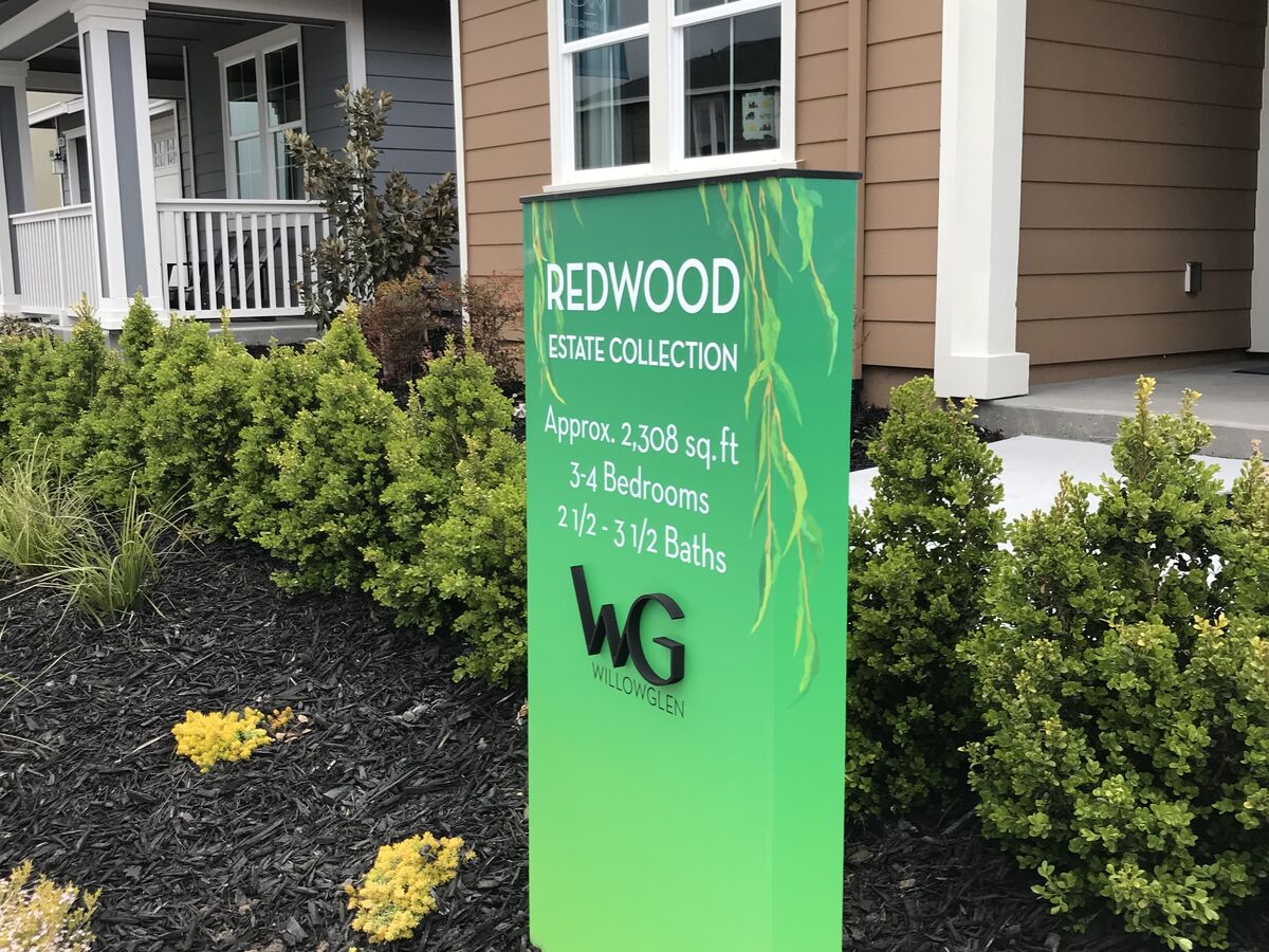 Green freestanding marker sign in a planting bed outside a new home, reading Redwood Estate Collection with square footage, bedroom and bathroom counts and the WillowGlen monogram