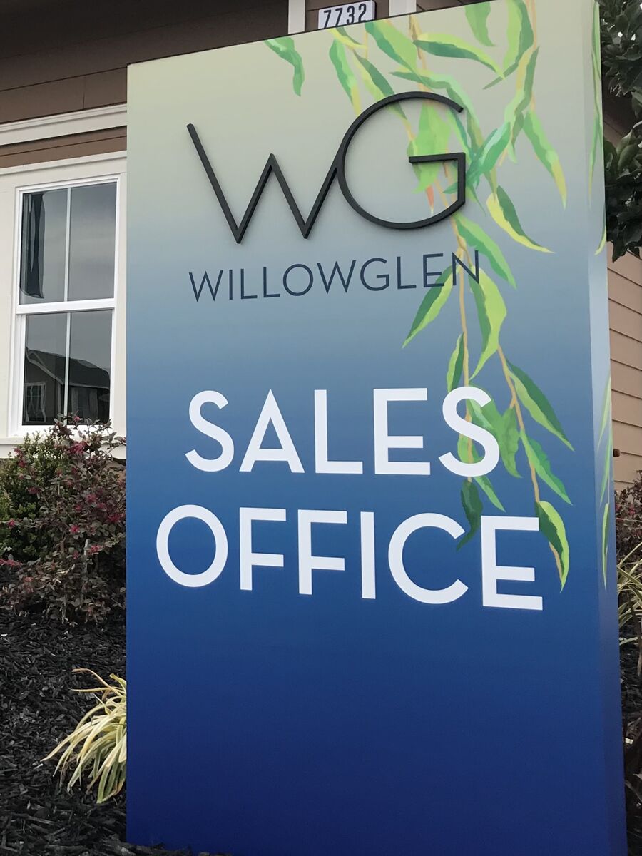 Freestanding panel sign outside a house, its face a blue-to-cream gradient with a painted willow branch, carrying dimensional WG letters above the word WillowGlen and, below, Sales Office in large white type